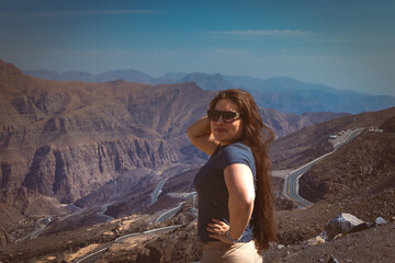 Fototapeta premium Portrait of a happy woman hiker standing on the slope of mountain ridge against mountains, blue cloudy sky on background. The woman is happy and looking at camera. Travel and active lifestyle concept.