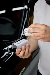 Specialistic ceramic coating liquid poured onto sponge by unrecognizable professional in protective gloves. Car detailing process. Black car in the background.