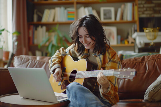Young Woman Learning Guitar Using Laptop. A young woman smiles as she plays guitar, using her laptop for an online music lesson in a cozy home setting.