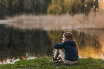 Little beautiful red-haired girl, lonely dreaming child sits on green grass against the backdrop of a lake in nature outdoors on vacation in the countryside. Photography, portrait, lifestyle.