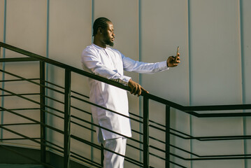 Happy young African American man in dashiki ethnic clothes taking selfie on wall and stairs background copy space. Millennial generation student and youth.