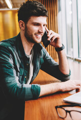 Good looking cheerful smart IT programmer communicating with friends using cellular connected to 4G internet.Successful hipster guy talking on telephone during installing application on laptop