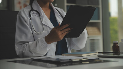 Serious female doctor using laptop and writing notes in medical journal sitting at desk. Young woman professional medic physician wearing white coat and stethoscope working on computer at workplace.
