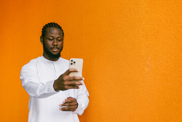 Happy young African American man in dashiki ethnic clothes taking selfie on orange wall background...