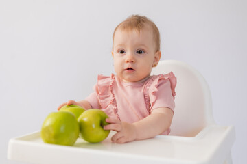 Fototapeta premium Cute baby in a baby chair nibbles apples on a white background