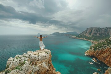 woman standing on rocks overlooking the sea, arms outstretched, yoga pose, mediterranean coast in background, cloudy sky - Powered by Adobe