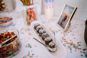 Valmiera, Latvia - August 19, 2023 - A close-up of a candy bar setup with various sweets, featuring a bowl of gummies, a plate of chocolate-coated coconut balls, and festive confetti on the table.