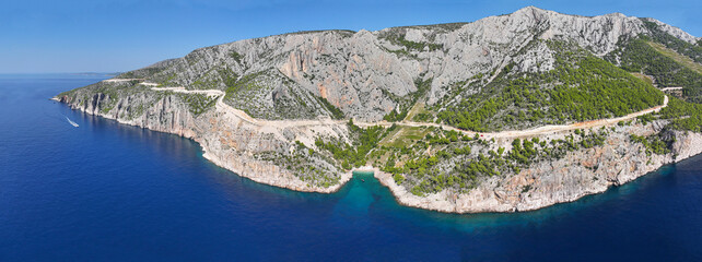 AERIAL Winding road cuts underneath the rocky cliffs and along the turquoise sea - obrazy, fototapety, plakaty