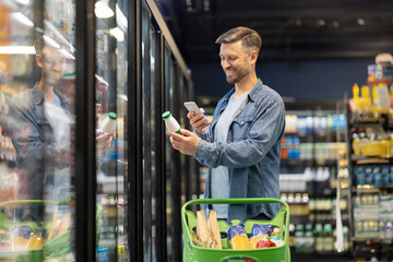 Smiling man choosing dairy products in supermarket, holding bottle and smartphone, scanning bar code on product through mobile phone