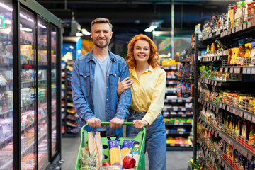 Happy family husband and wife walking with shopping cart full of grocery, shopping together at supermarket, smiling at camera