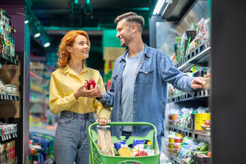 Happy European couple on grocery shopping, choosing fresh vegetables, standing with trolley cart in supermarket, purchasing essentials