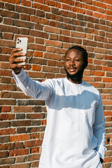 Happy young African American man in dashiki ethnic clothes taking selfie on brick wall background....