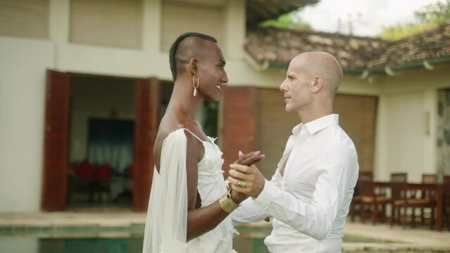 Multiethnic androgynous gay couple dance waltz on outdoor wedding ceremony at luxury villa on tropical island. LGBTQIA biethnic men at trendy fashionable engagement session. Same sex marriage union.