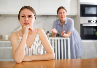 Offended young girl sitting at the kitchen table, mother standing behind and trying to talk to her...