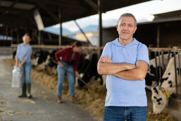 Positive middle-aged man farmer standing in cowshed with arms crossed and smiling.
