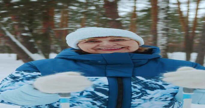 Vertical video. Portrait of a happy elderly woman who looks at the camera with a positive smile while doing winter sports in a snowy forest. Sports pensioners go skiing on classic skis. 4k footage
