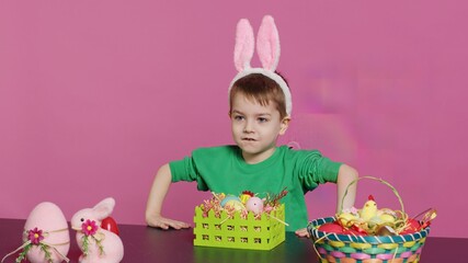 Obraz premium Sweet young boy making colorful arrangements for Easter holiday festivity, putting painted eggs in a handcrafted basket. Ecstatic joyful kid using crafting materials to create decorations. Camera A.