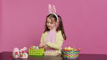 Obraz premium Smiling happy schoolgirl presenting a handmade decorated basket in studio, making easter holiday preparations against pink background. Young child showing handcrafted arrangements. Camera A.
