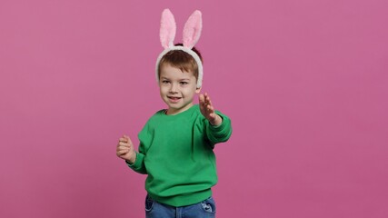 Obraz premium Cute little boy wearing fluffy bunny ears in studio and waving, being adorable against pink background. Smiling young kid being excited about easter holiday festivity, childhood innocence. Camera A.