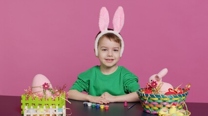 Obraz premium Young small kid placing bunny ears on his head in studio, preparing for easter sunday holiday celebration. Cheerful little boy sitting at a table to paint eggs and craft festive ornaments. Camera A.