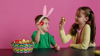 Obraz premium Sweet children knocking eggs together for easter tradition in studio, playing a seasonal holiday game against pink background. Lovely adorable kids having fun with festive decorations. Camera B.