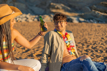  Hispanic woman offers a beer to her boyfriend as they sit together on the sandy beach during a...