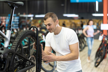 Young male customer choosing bike and checking tire on bicycle wheel in modern sport store