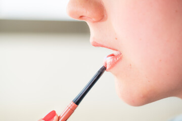 A young teenager paints her lips with a small brush of light pink gloss. She is reflected in a...