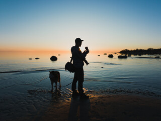 Male drone pilot and photographer with husky dog ​​on the beach at sunset.