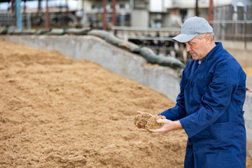 Portrait of positive man farmer in uniform checking beer oilcake in fodder storage