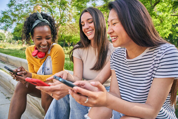 Three young women are sharing happy moments on a bench, smiling at their cell phones. They sit...