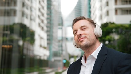Caucasian businessman listen headphone and dancing to relaxing music with cheerful and motivation. Skilled project manager enjoy to listen relaxed song with blurred background at urban city. Urbane.