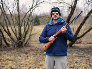 Man holding small caliber rifle for shooting practice in a country side. Sport and entertainment. Male with grey beard in blue jacket and dark hat. Out door activity.
