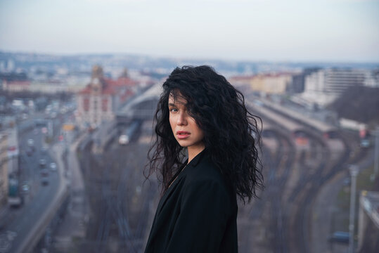 Attractive young brunette woman in a black jacket stands on a rooftop against a city space background.