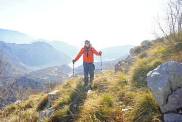 Obraz premium A tourist in a bright jacket with trekking poles poses against the backdrop of a picturesque mountain landscape. Hiking on Mount Orjen in Montenegro