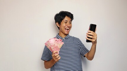 young asian man happy posing holding money and looking at smartphone
