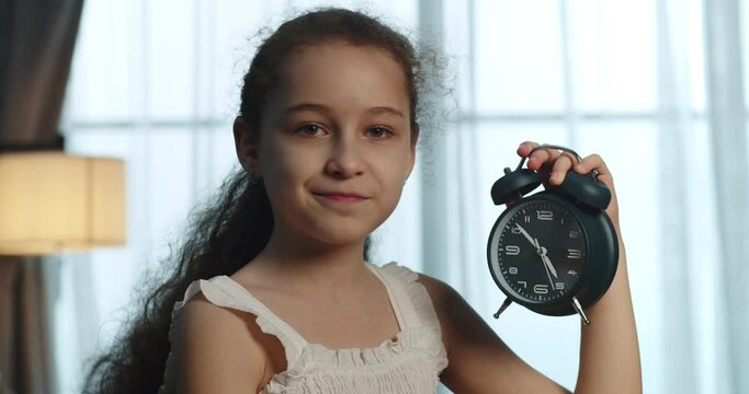 A happy cute little girl in a white dress holds an alarm clock in her hands, the alarm clock rings, the child makes an anxious and joyful face and says the time. Educational lifestyle. 4K