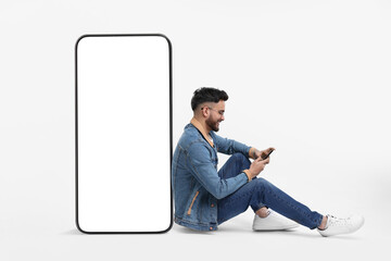 Man with mobile phone sitting near huge device with empty screen on white background. Mockup for...
