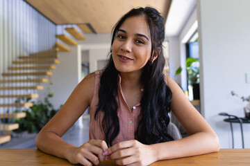 Young biracial woman sitting at table, smiling at camera on a video call at home