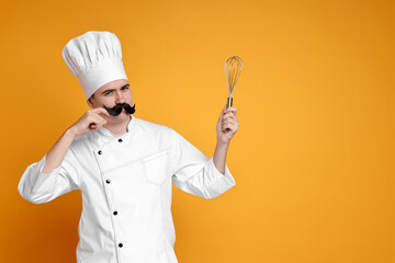Portrait of happy confectioner with funny artificial moustache holding whisk on orange background,...
