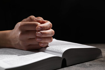 Religion. Christian woman praying over Bible at table against black background, closeup. Space for text