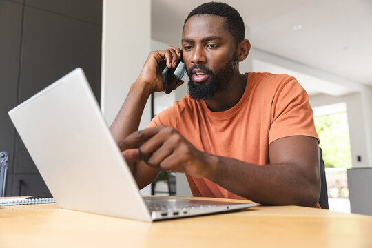 African American male homeowner talking on phone, using laptop at home