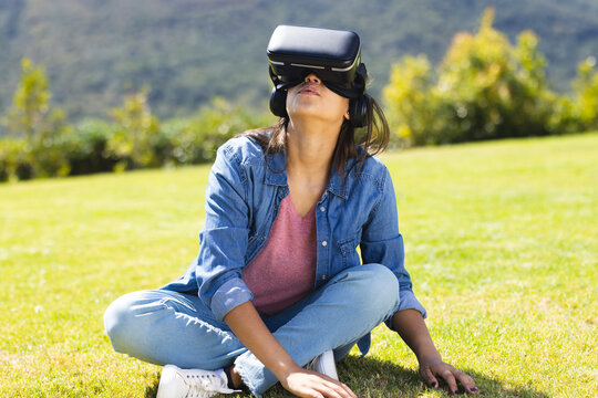A young biracial woman wearing VR headset outdoors, relaxing on grass