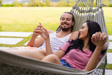 Diverse couple relaxing outdoors in hammock, man holding smartphone
