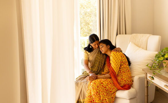 Indian mother comforting teenage daughter on sofa, both in traditional attire, copy space
