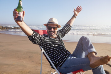 happy mature man sitting on the beach with arms up enjoying a beer and solitude in retirement 