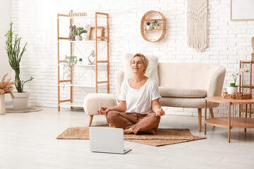 Mature woman with laptop meditating at home. Online yoga classes