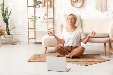 Mature woman with laptop meditating at home. Online yoga classes