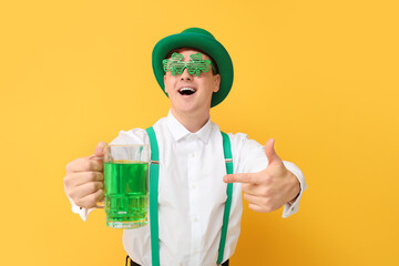 Happy young man in leprechaun's hat with party glasses pointing at mug of beer on yellow...