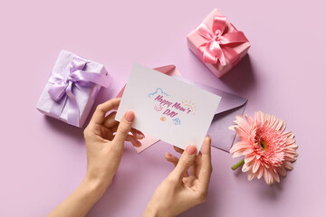 Female hands with festive postcard, gift boxes and flower on lilac background. Happy Mother's Day